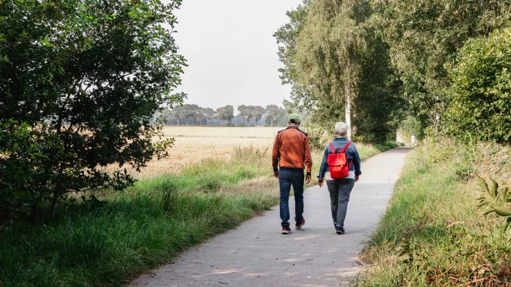Ouder stel met rugzak wandelt over een fietspad langs de akkers