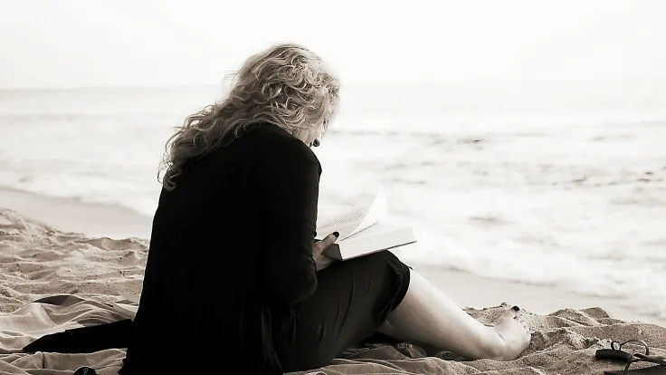 Vrouw zit met een boek op een kleed in het zand met uitzicht op zee