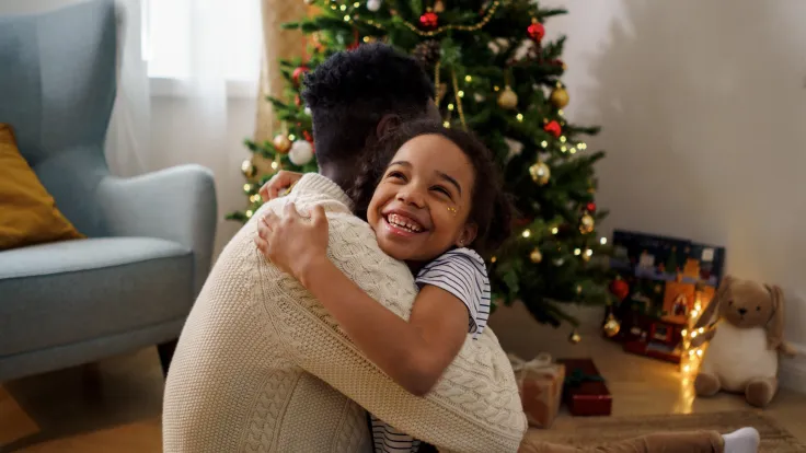 Kind knuffelt lachend een volwassene voor de versierde kerstboom